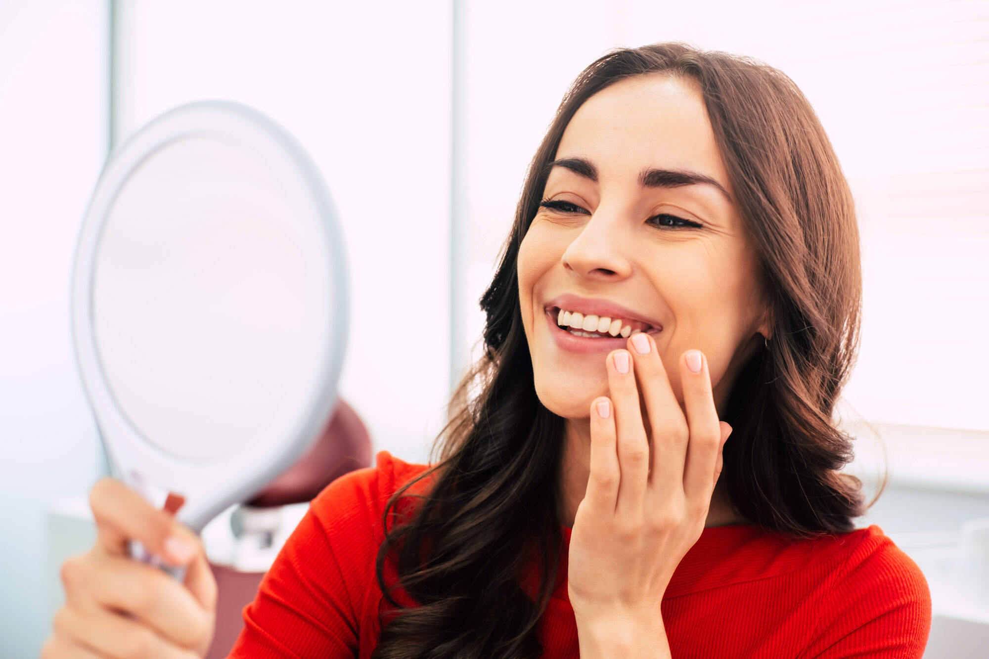Honed to perfection. Fabulous woman clothed in red sweater, with curly long hair and elegant hands is holding a mirror for looking through the final work of dental doctor. Dental Implants